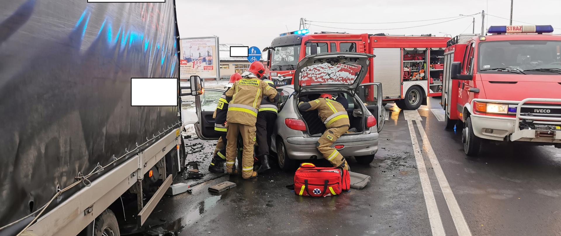 Zdjęcie przedstawia 4 strażaków podczas wypadku, próbujących dostać się do osoby poszkodowanej w pojeździe, na drodze stoi samochód dostawczy i osobowy a w tle dwa auta bojowe straży pożarnej