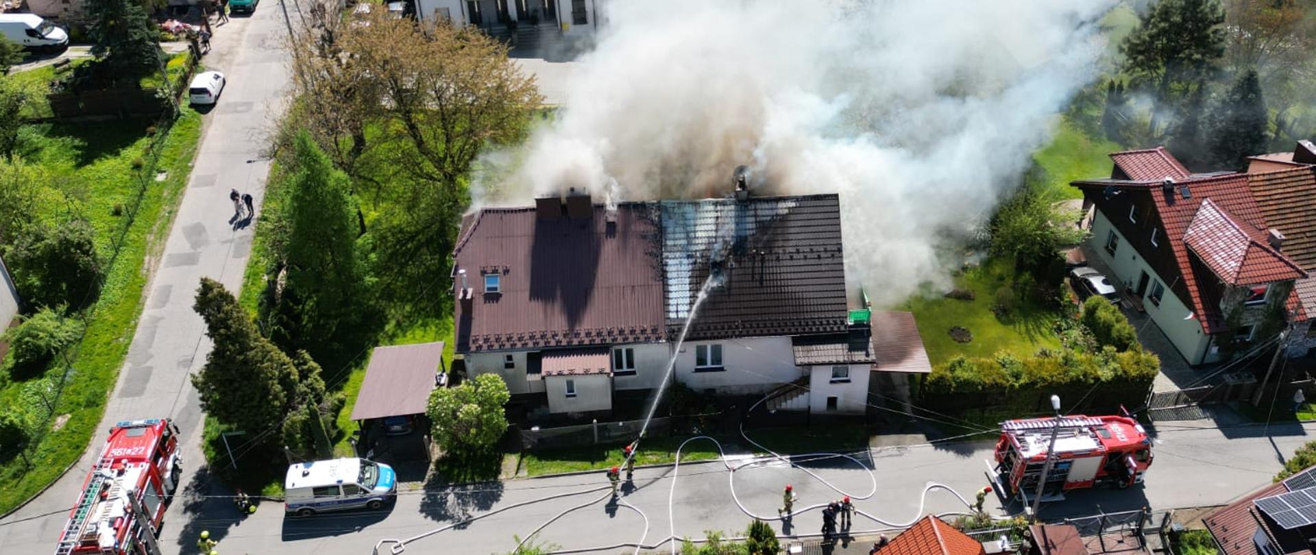 Na zdjęciu palący się budynek mieszkalny, widać wydobywający się dym. W pobliżu dwa samochody pożarnicze oraz strażacy prowadzący działania gaśnicze