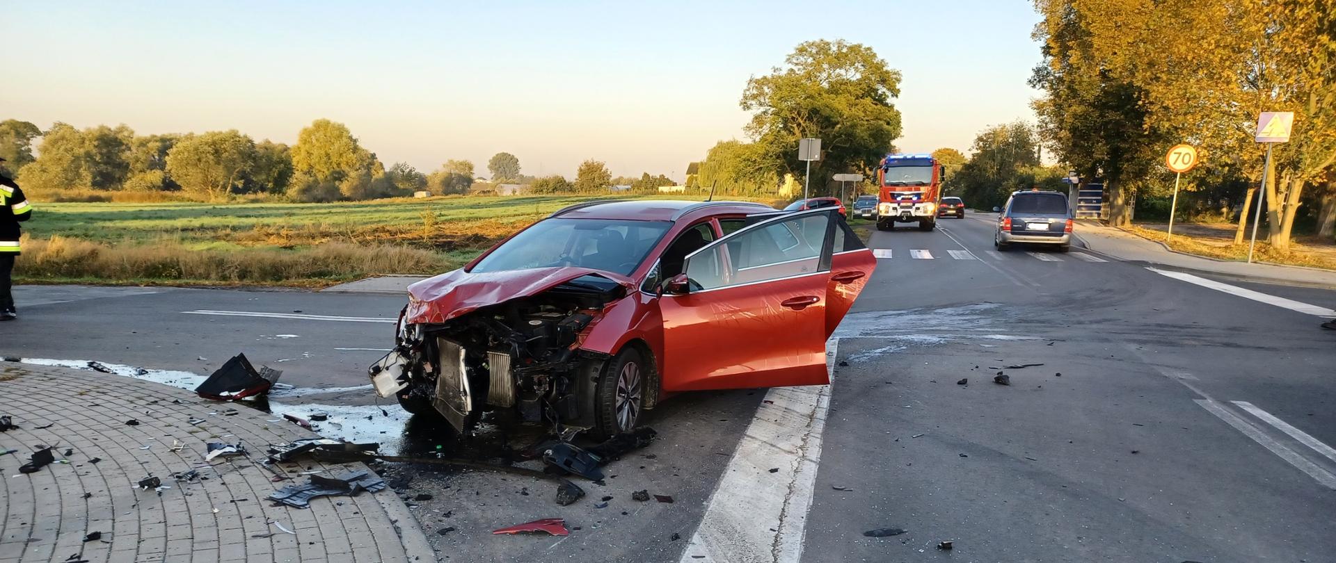 Zdjęcie obrazuje drogę asfaltową koloru ciemno szarego z namalowanymi znakami poziomymi na niej (zerba –przejście dla pieszych). W dolnej lewej części zdjęcia widać chodnik z ułożonych kostek typu polbruk. W tle widzimy drzewa liściaste koloru zielonego, przydrożne znaki drogowe oraz przystanek autobusowy, wiata w kolorze niebiesko srebrnym. Na pierwszy plan zdjęcia wysuwa się uszkodzony samochód osobowy marki KIA koloru bordowego. Samochód ma otworzone tylne drzwi pasażera oraz przednie drzwi od strony kierowcy. Uszkodzony pojazd stoi przodem do zdjęcia, gdzie widać uszkodzoną maskę pojazdu oraz brak przedniego zderzaka. Widać pod maską całą komorę silnika wraz z chłodnicą. W pojeździe nikt nie przebywa. Na drodze wokół pojazdu są porozrzucane różne, trudne do zidentyfikowania elementy karoserii oraz części pojazdu. Widać również wyrwany przedni reflektor uszkodzonego samochodu, który leży na drodze w dolnej prawej części zdjęcia.