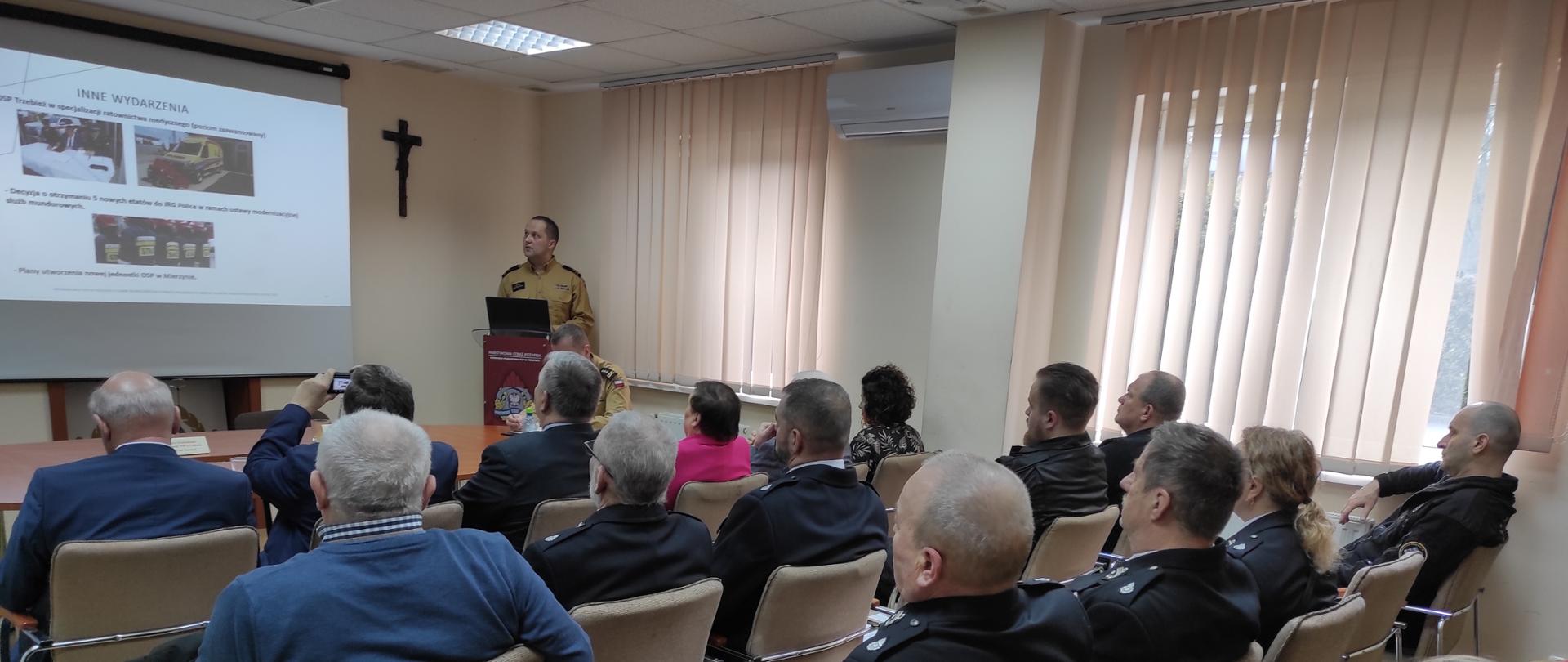 Na zdjęciu widać fragment wnętrza pomieszczenia z ekranem, na którym wyświetlana jest multimedialna prezentacja. Obok ekranu, po prawej stronie krzyż. Obok mężczyzna w piaskowym mundurze. Na pierwszym planie widoczne plecy zaproszonych gości, siedzących na jasnych krzesłach ustawionych w szeregi.
