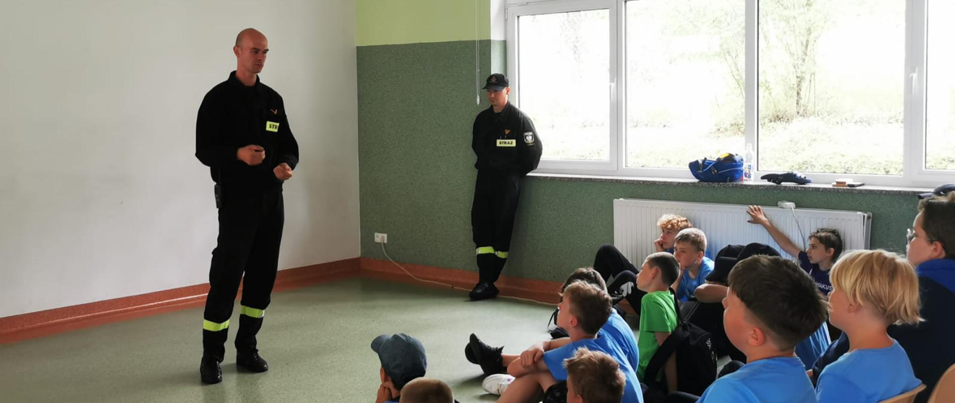 Zdjęcie przedstawia dwóch strażaków w szkolę podczas zajęć edukacyjnych na sali. młodzież siedzi i słucha strażaków.