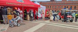 namiot Państwowej Straży Pożarnej na dziedzińcu rynku Manufaktury, widoczni ludzie oglądający wystawkę, obok namiotu kartonowa postać strażaka przy której ludzie robią sobie zdjęcia, stoi samochód elektryczny Melex z napisem straż oraz z lampami błyskowymi na dachu, dzieci przebrane w strażaków wsiadają do samochodu elektrycznego, w tle budynek z czerwonej cegły