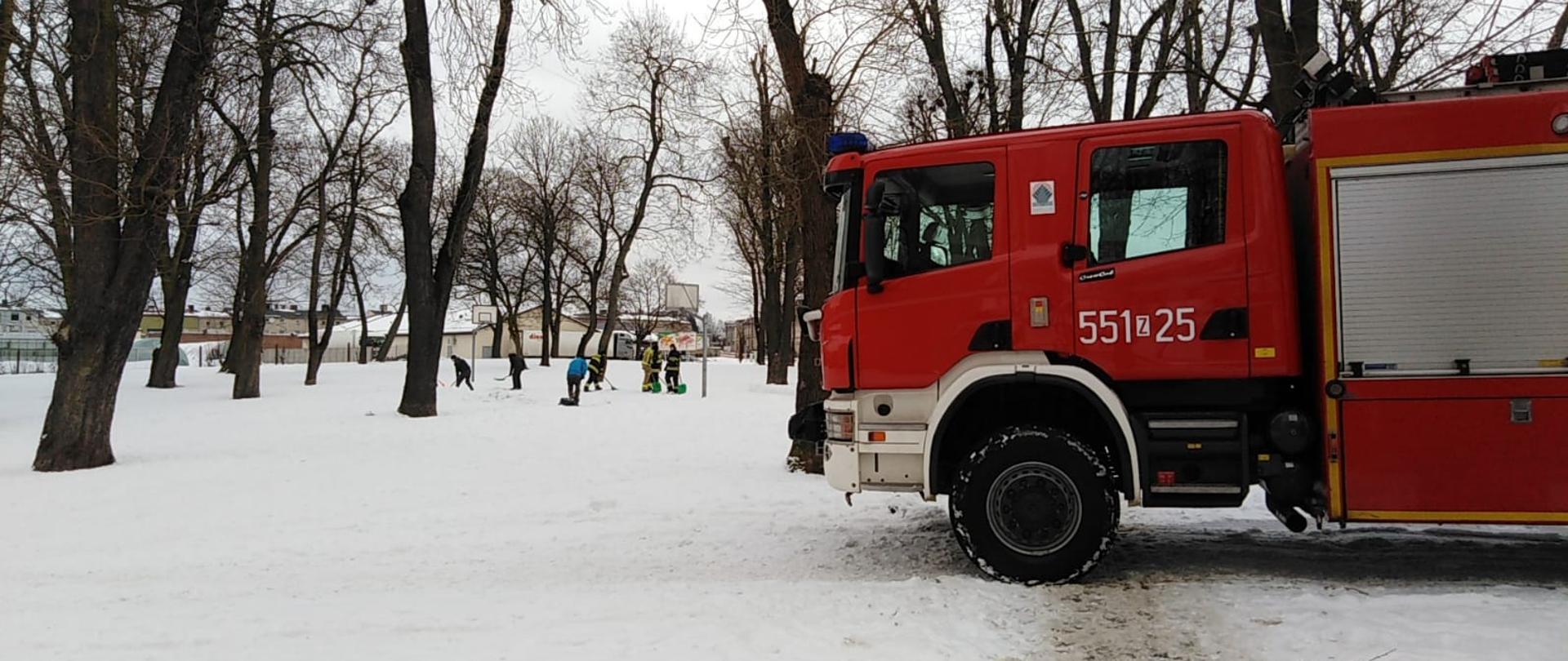 Zdjęcie przedstawia samochód strażacki stojący w pełnym śniegu parku z drzewami. Na aucie widnieje numer pojazdu 551z25. W tle widać odśnieżające osoby oraz budynki.