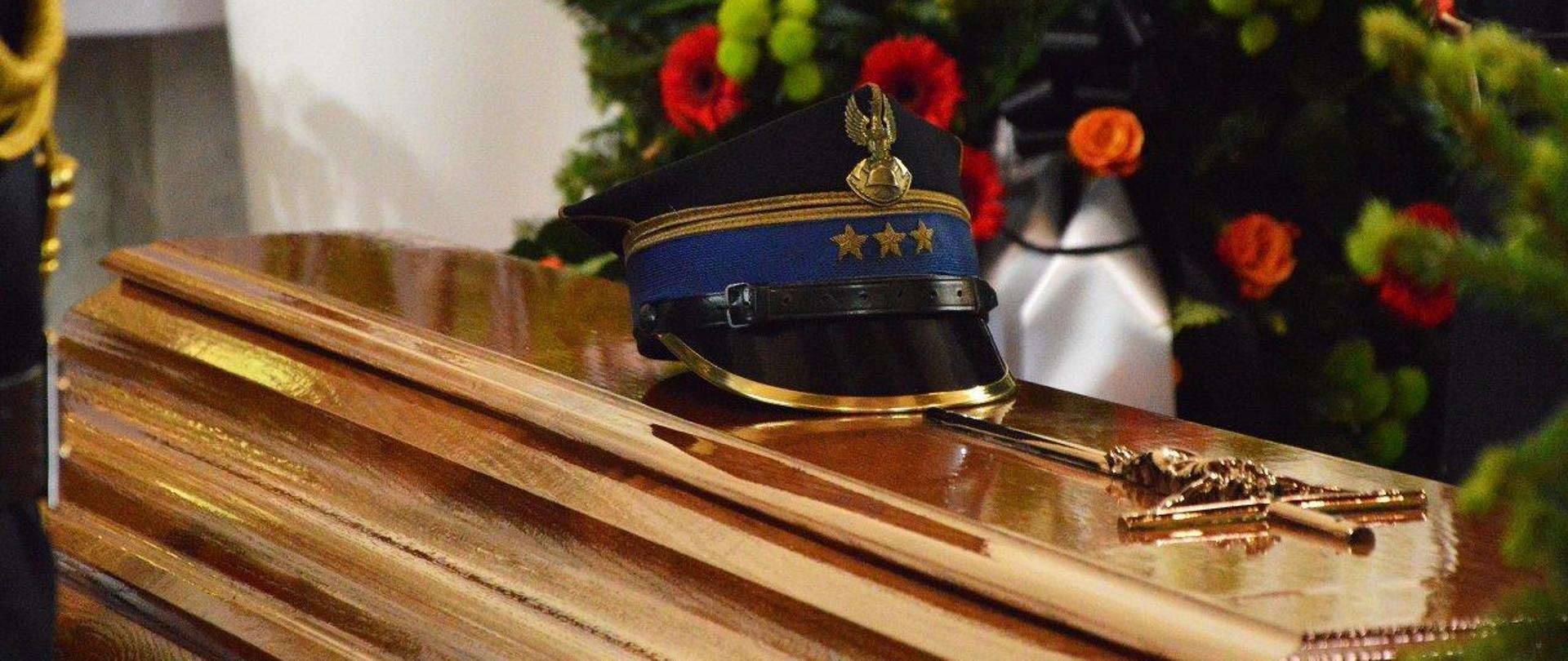 Trumna w kolorze brązowym, ze złotym krzyżem. Na trumnie leży czapka oficera pożarnictwa. W tle wieńce pogrzebowe.