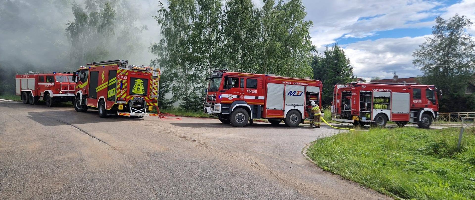 Na zdjęciu cztery samochody strażackie podczas działań gaśniczych, u góry z lewej strony unosi się dym. W tle zielone drzewa.