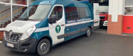 Zdjęcie przedstawia samochód Inspekcji Transportu Drogowego. Samochód znajduje się na placu przed garażami jednostki ratowniczo-gaśniczej. Brama od garażu jest otwarta. W garażu znajduje się wóz strażacki