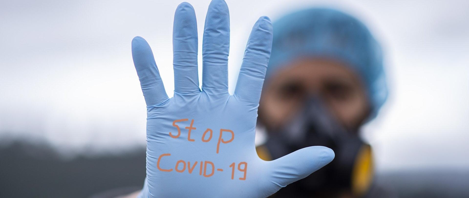 Na pierwszym planie ręka w jednorazowej rękawiczce, na której jest napisane Stop Covid-19, a którą wystawił człowiek stojący na drugim planie nieostry w maseczce i z czepkiem medycznym na głowie.
