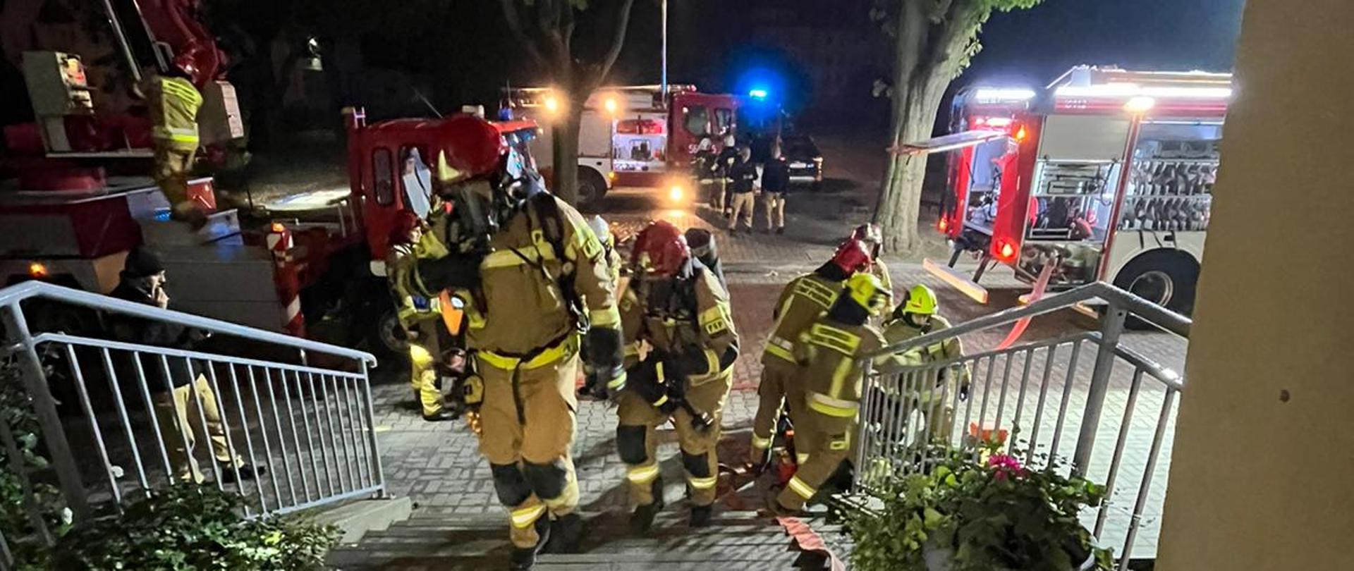 Dwóch strażaków w ubraniach specjalnych, hełmach i aparatach ochrony dróg oddechowych idzie po schodach, za nimi widoczne są wozy gaśnicze i ćwiczący strażacy