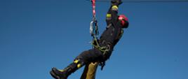Widok z dołu. Funkcjonariusz PSP przemieszczający się po linie wyciągu z wykorzystaniem metod alpinistycznych. W tle błękitne niebo. 