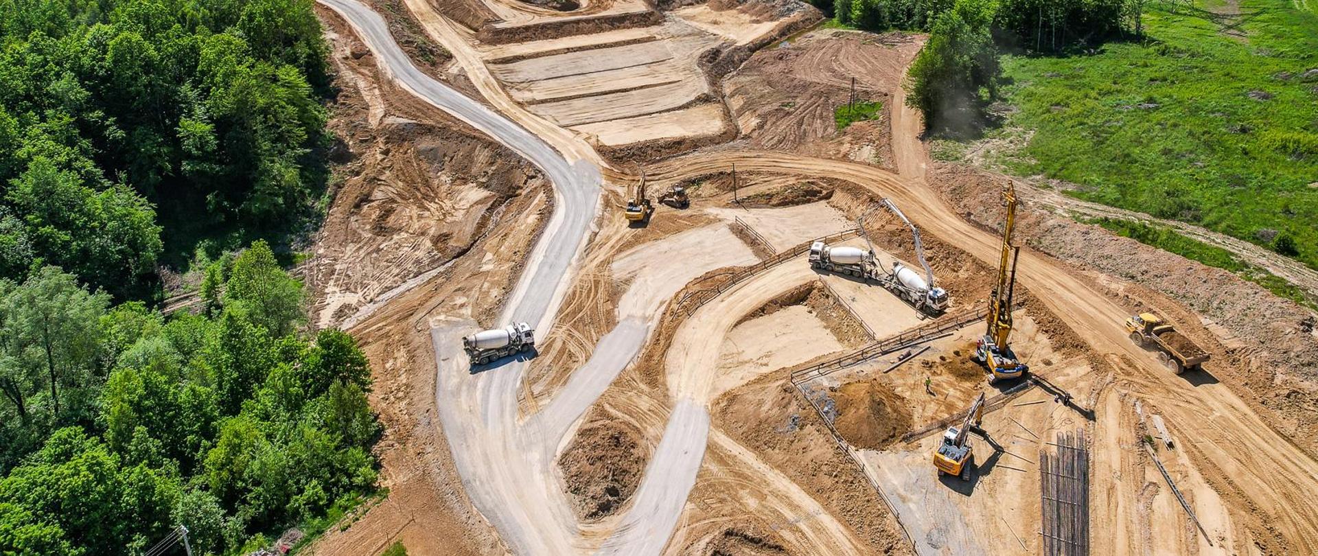 Widok z góry na budowę S19 Babica - Jawornik. Pogoda słoneczna, świeci słońce. Wśród drzew widać wytyczoną trasę przyszłej drogi oraz pracujący sprzęt budowlany - palownice, koparki, betoniarki.
