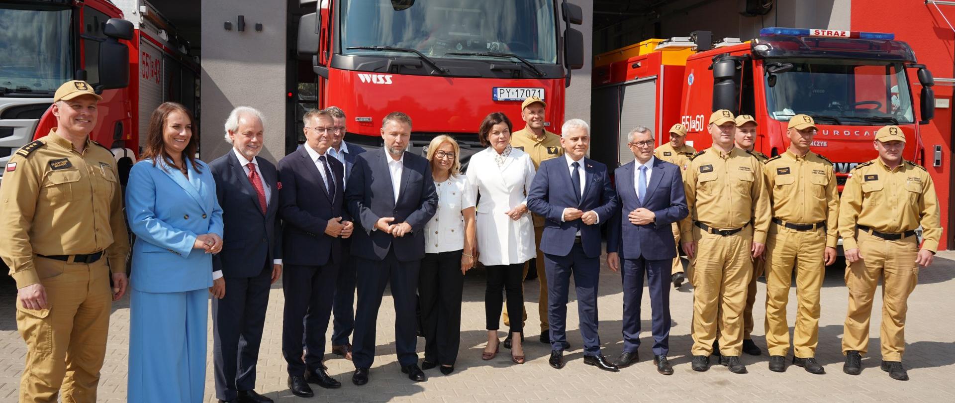 Grupa strażaków w piaskowych mundurach oraz osób w strojach formalnych pozuje do wspólnego zdjęcia przed remizą strażacką. W tle stoją trzy czerwone wozy strażackie, częściowo wysunięte z otwartych bram garażowych, a fotografia została wykonana w słoneczny dzień podczas oficjalnego wydarzenia.