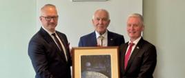 3 mężczyzn, w tym wojewoda, w oficjalnych ubraniach trzyma w dłoniach obraz, pozują do zdjęcia