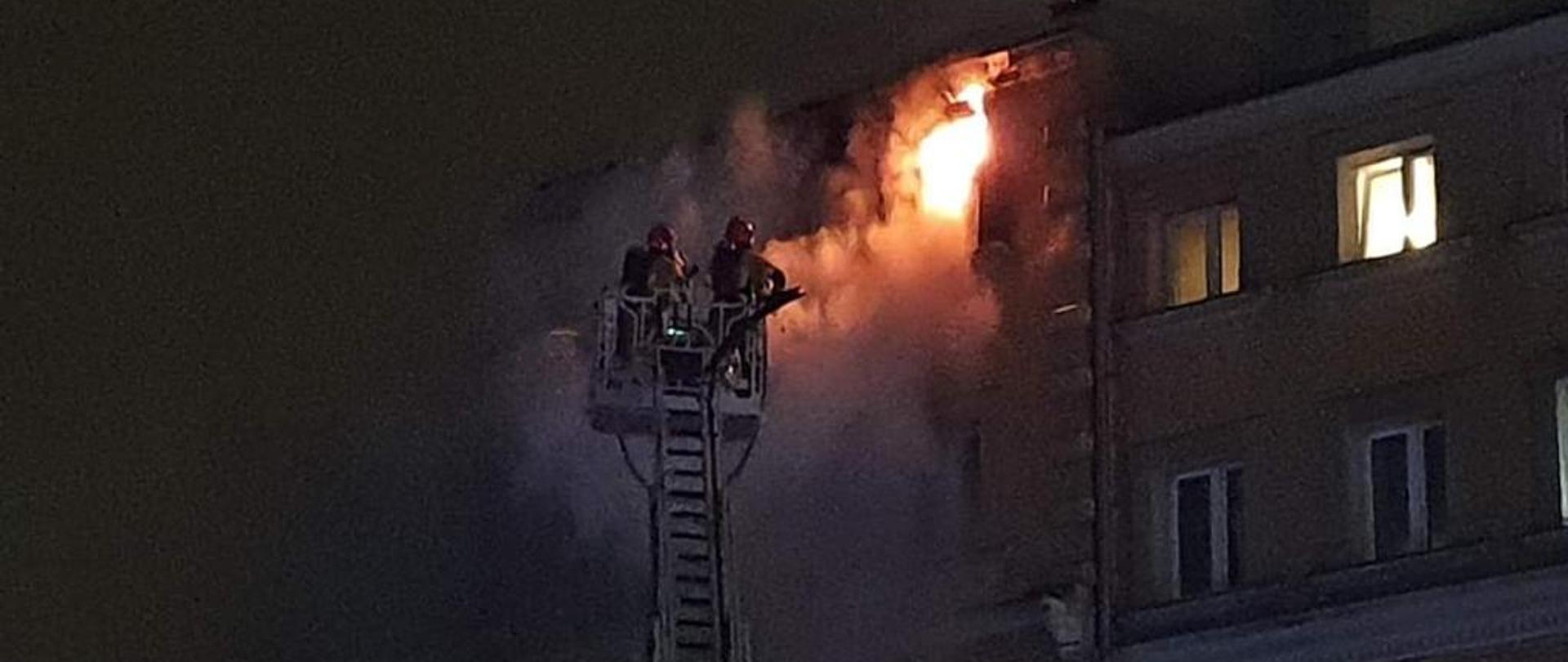 Na zdjęciu widać budynek, z jednego okna na ostatniej kondygnacji wydobywają się płomienie. Przed budynkiem widać wysunięta drabinę mechaniczną z koszem w którym znajduje się dwóch strażaków