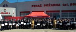 Dzień. Widok na budynwk o białej elewacji na której znajdują się napisy w kolorze czerwonym "Straż Pożarna tel. 998: i "JRG nr 1" . W elewacji znajdują się 4 bramy wjazdowe w kolorze czerwonym z przeszkleniami. Na tle budynku stoi duża grupa osób w większości ubranych w mundury wyjściowe OSP. Wszyscy pozują do zdjęcia