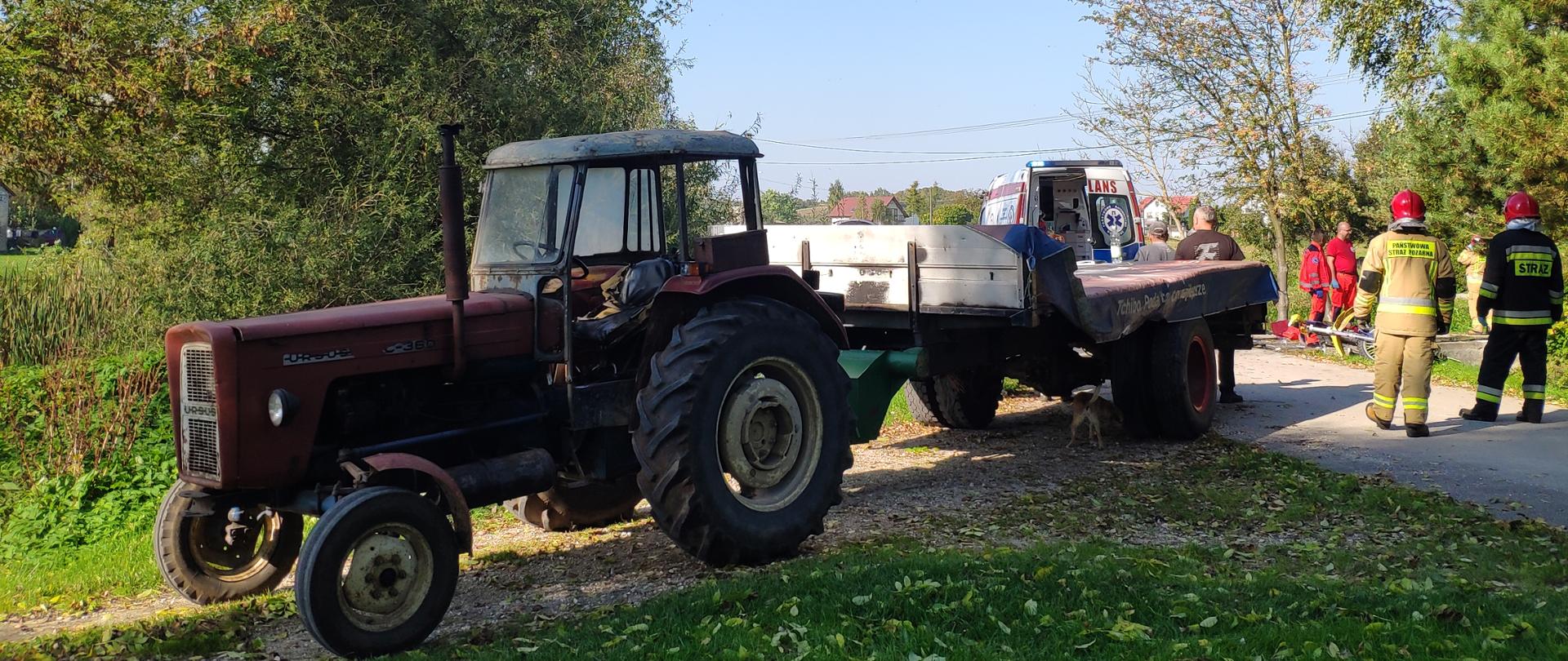 Zdjęcie przedstawia ciągnik z przyczepą stojący na drodze. Za nim z prawej strony w oddali widać przewrócony motocykl oraz strażaków i ratowników z Pogotowia Ratunkowego.

