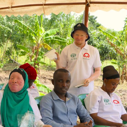 Grupa osób, część z koszulkach z napisame Adra/Polish Aid siedzi na krzesłach, dwie osoby są Albinosami