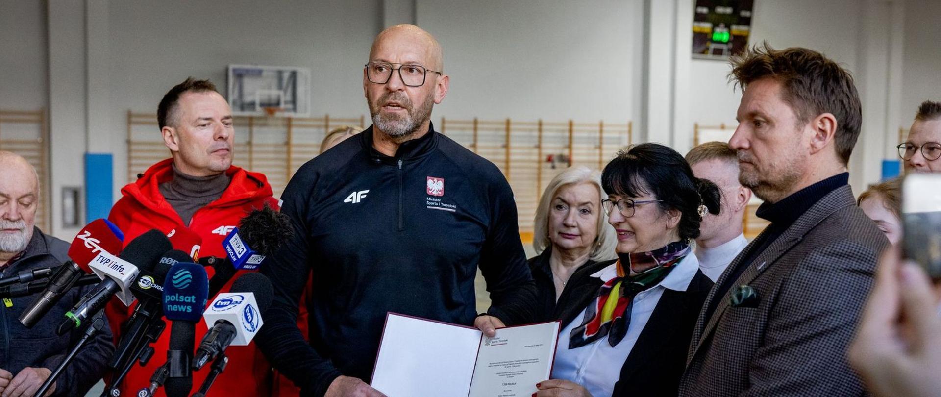 Konferencja prasowa. Minister sportu i turystyki Jakub Rutnicki trzyma w rękach decyzje o dofinansowaniu budowy toru saneczkowego w Karpaczu. Z lewej strony stoi sekretarz stanu Piotr Borys. Po bokach inni uczestnicy konferencji. Na pierwszym planie widać mikrofony, w tle drabinki gimnastyczne.