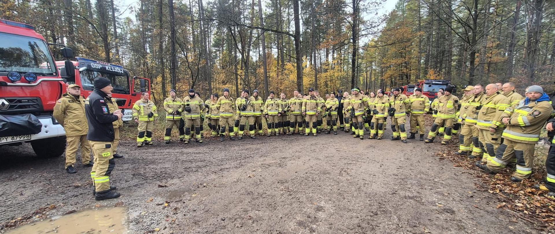 Zdjęcie przedstawia dużą grupę strażaków zgromadzonych na odprawie w terenie leśnym, prawdopodobnie po zakończeniu ćwiczeń lub akcji gaśniczej.
• Na pierwszym planie znajduje się szeroka, błotnista i mokra droga gruntowa, z kałużami (szczególnie widocznymi po lewej stronie).
• W centrum stoi duża grupa, licząca około 30-40 strażaków, ubranych w typowe, żółto-piaskowe mundury bojowe z odblaskowymi pasami.
o Są ustawieni w półkolu lub luźnym szyku.
o Wszyscy strażacy mają poważny wyraz twarzy, co sugeruje skupienie na treści odprawy.
• W lewej części kadru dwóch mężczyzn, z których jeden jest prawdopodobnie dowódcą lub instruktorem, zwraca się do grupy. Mężczyzna po lewej jest ubrany w ciemniejszą (czarną/granatową) kurtkę, co może odróżniać jego stopień lub funkcję od reszty.
• Tło stanowią liczne wozy strażackie, co potwierdza charakter spotkania.
o Po lewej widoczny jest przód czerwonego wozu (prawdopodobnie Renault, sądząc po kształcie lamp) z częściowo widoczną tablicą rejestracyjną WPY 47998.
o Za grupą, w głębi lasu, widać co najmniej trzy kolejne czerwone pojazdy, w tym jeden z napisem OSP KLWÓW.
• Otoczenie to las liściasty i mieszany, na co wskazują pnie drzew i jesienne barwy ulistnienia (żółcie i brązy) widoczne za strażakami.
Ogólne wrażenie: Scena przedstawia zorganizowaną przerwę, podsumowanie działań lub wydawanie poleceń dużej grupie ratowników, co podkreśla ich profesjonalizm i gotowość operacyjną w trudnych warunkach terenowych
