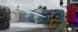 Akcja na składowisku odpadów. Plac, na którym znajdują się pryzmy z odpadami. Na pierwszym planie fragment wozu straży pożarnej oraz strażak gaszący pożar odpadów przy użyciu piany gaśniczej. Na ziemi znajduje się rozwinięty wąż strażacki koloru żółtego.