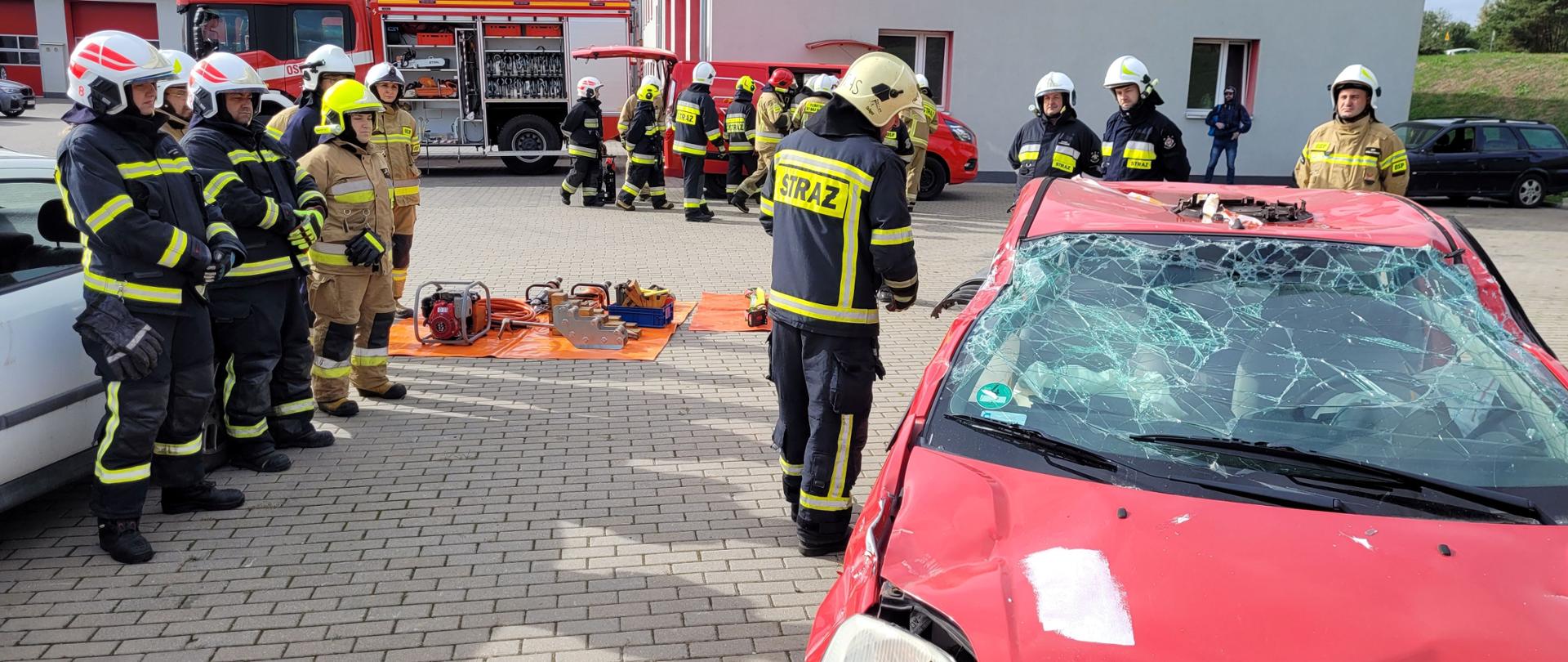 Strażacy w piaskowych i granatowych mundurach stojący w grupie i podczas szkolenia przy rozbitym aucie. W tle budynek z czerwonymi bramami wyjazdowymi i pojazdy pożarnicze..