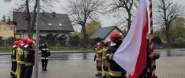 Funkcjonariusze stojący w umundurowaniu bojowym na uroczystej zbiórce. Trzej z nich stoją przy maszcie i podnoszą flagę państwową.