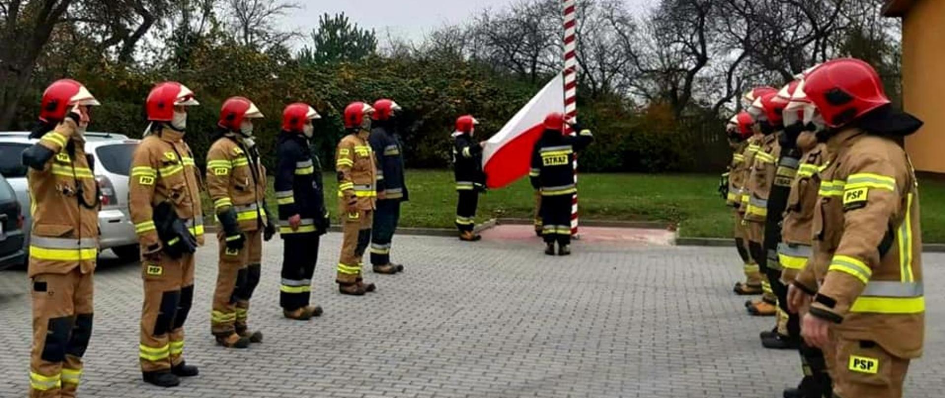 Zdjęcie przedstawia funkcjonariuszy JRG2 podczas wciągania flagi z okazji uroczystości Święta Odzyskania Niepodległości, ubranych w uranie bojowe, zabezpieczonych w maseczki.
