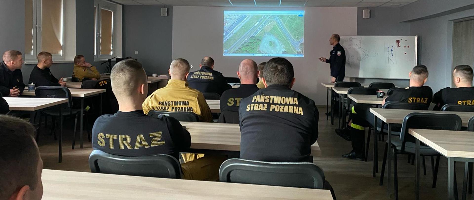 Jasne pomieszczenie, w którym przy stolikach siedzą strażacy w piaskowych i czarnych mundurach słuchając wykładu prowadzonego przez policjanta. W tle na ścianie wyświetlana prezentacja multimedialna.