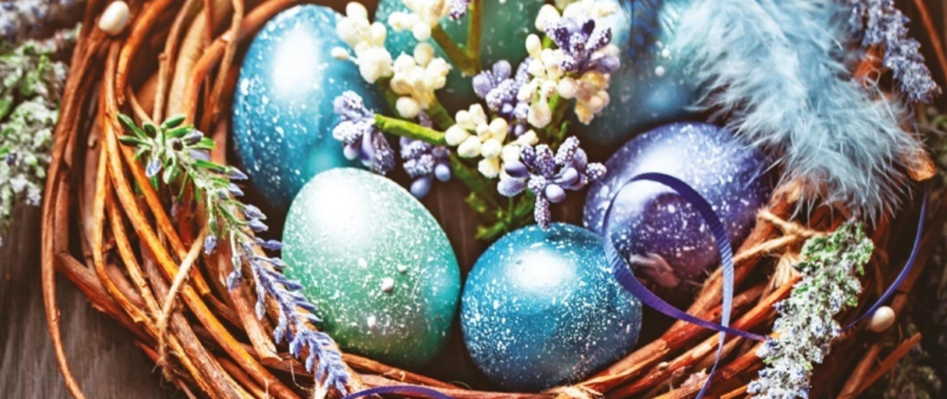 Zdjęcie przedstawia pisanki w wiklinowym koszyczku. W koszyczku jest 6 pisanek, 2 zielone, 3 niebieskie i jedna fioletowa, każda z nich ma duże ilości drobnych nieregularnych białych prążków. W środku koszyczka pomiędzy jajkami umieszczono biało-fioletowe kwiatuszki. W zewnętrznej części wikliny umieszczone gałązki fioletowych wrzosów oraz bazie. Koszyczek leży na drewnianym brązowym stole.