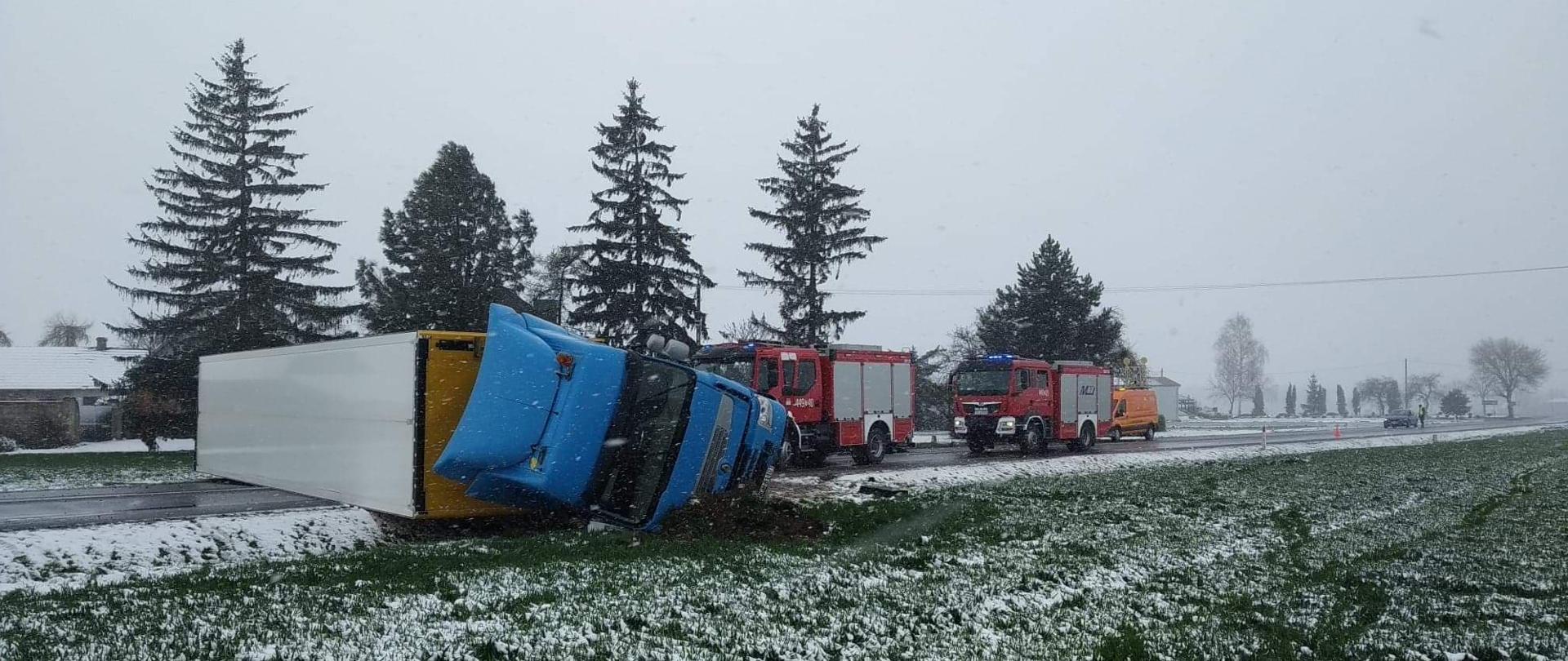 Widok na przewrócony na bok ciągnik siodłowy wraz z naczepą całkowicie blokujący przejazd na drodze, kabina ciągnika siodłowego poza jezdnią w rowie, naczepa całą długością na obu pasach drogi. W tyle za leżącym samochodem ciężarowym dwa samochody gaśnicze i samochód służby drogowej.