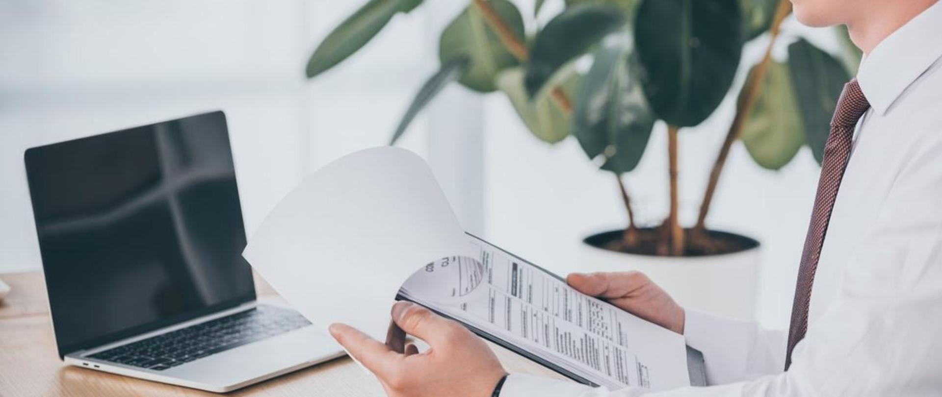 Z prawej strony widoczna część tułowia mężczyzny w białej koszuli trzymającego w rękach druk formularza, po lewej stronie na blacie biurka stoi otwarty laptop. W tle widoczna zielona roślinna w białej doniczce