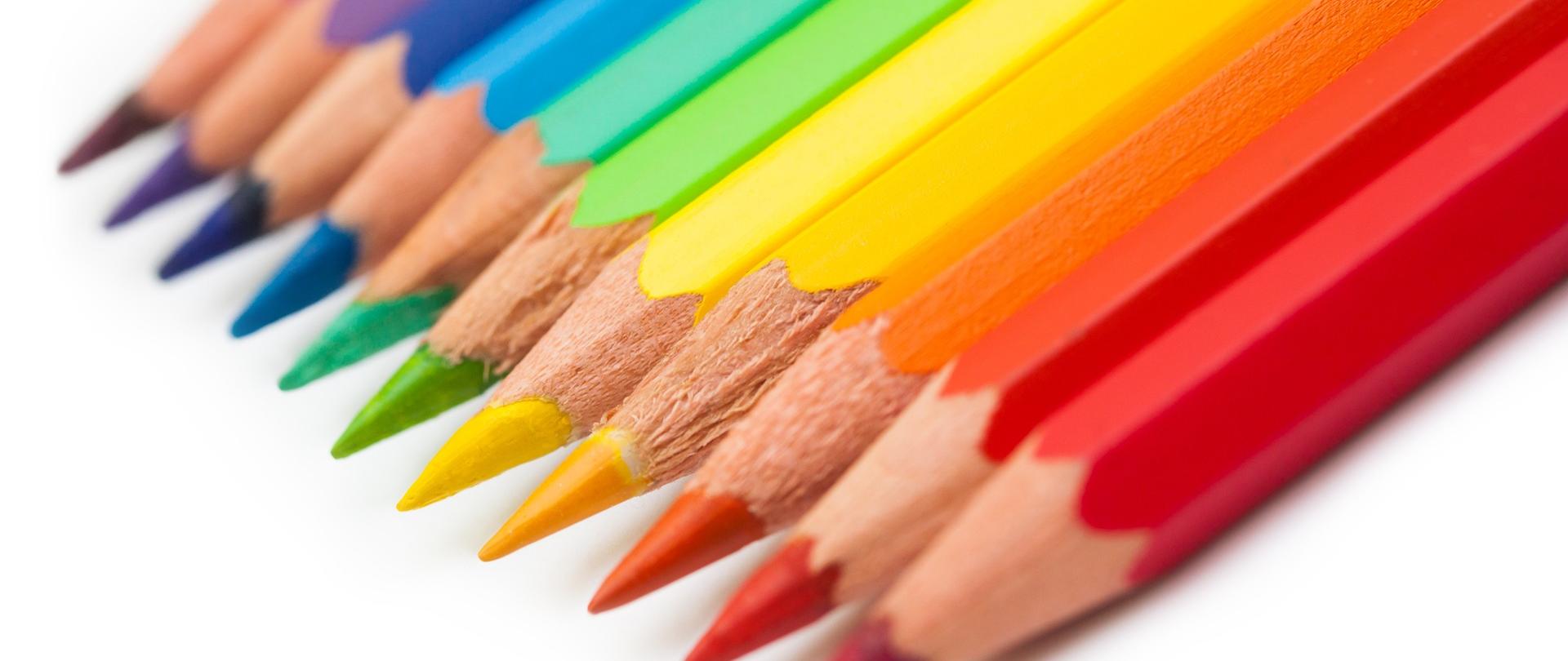 Zbliżenie na rząd ostro zakończonych kredek ołówkowych ułożonych w kolejności kolorów tęczy na białym tle. Kolory przechodzą od czerwonego, pomarańczowego, żółtego, zielonego, niebieskiego do fioletowego. Kredki są ułożone pod kątem, ich końcówki (grafity) są skierowane w lewo w górę i są w wyraźnej ostrości, podczas gdy reszta korpusów kredek staje się nieostra, dając efekt małej głębi ostrości. Końcówki kredek są świeżo zaostrzone.