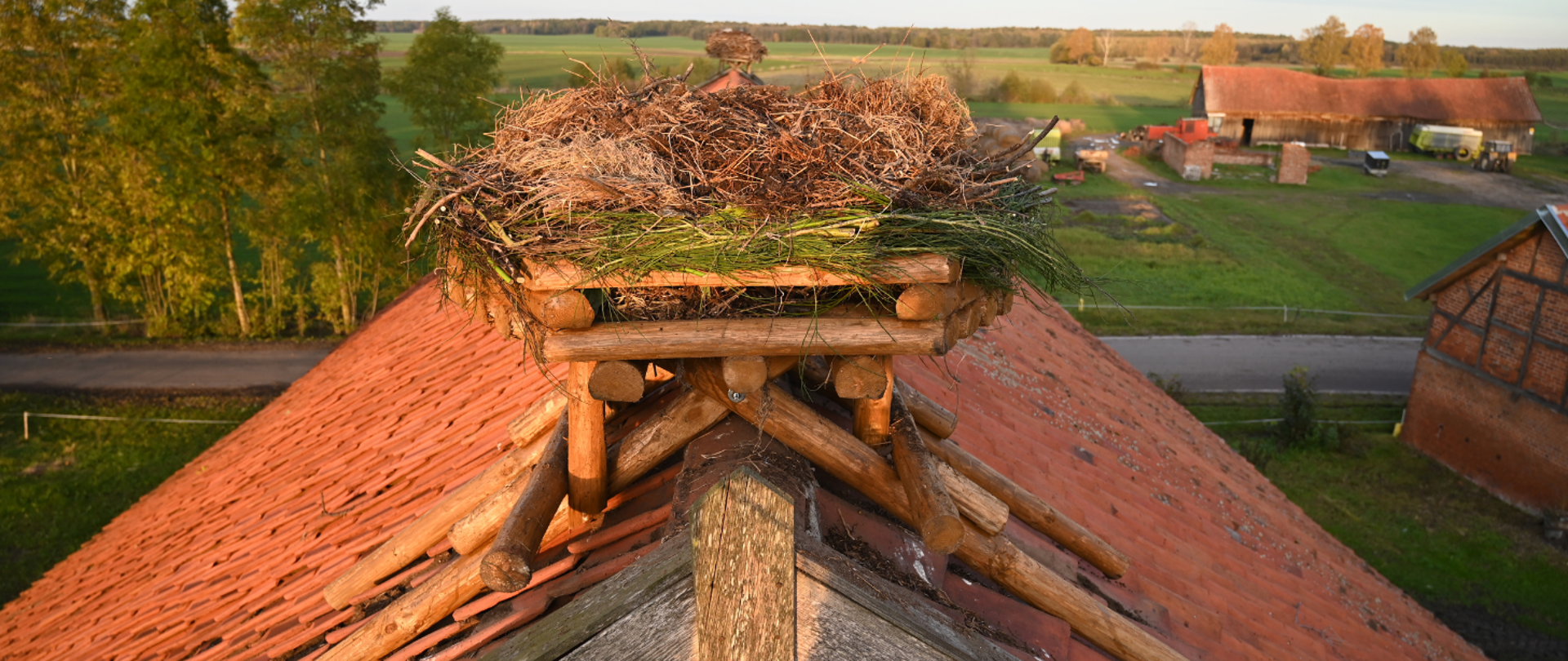 Gniazdo bocianie na dachu budynku z czerwonej dachówki. W oddali zabudowania i pola.
