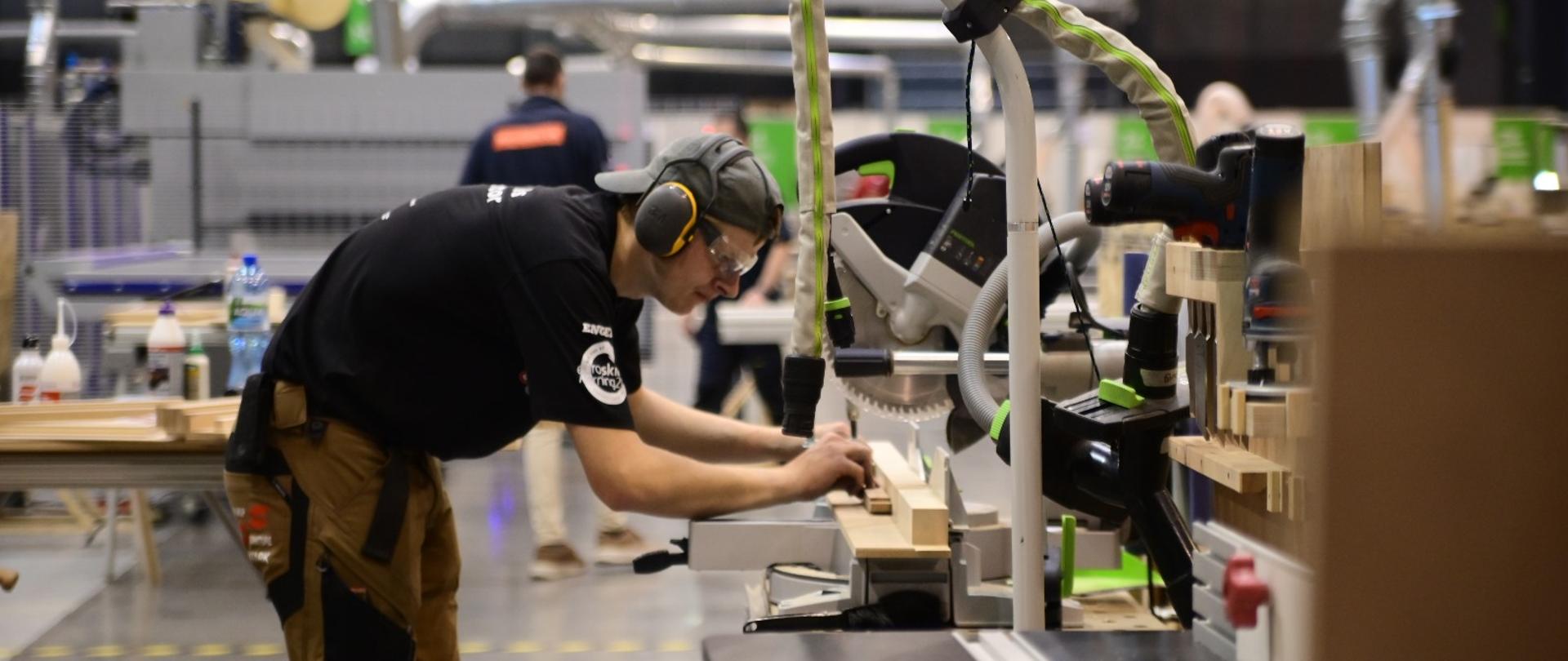 Mężczyzna w czarnej koszulce i ochronnikach słuchu i oczu pochyla się nad frezarką, która wycina drewnianą belkę.