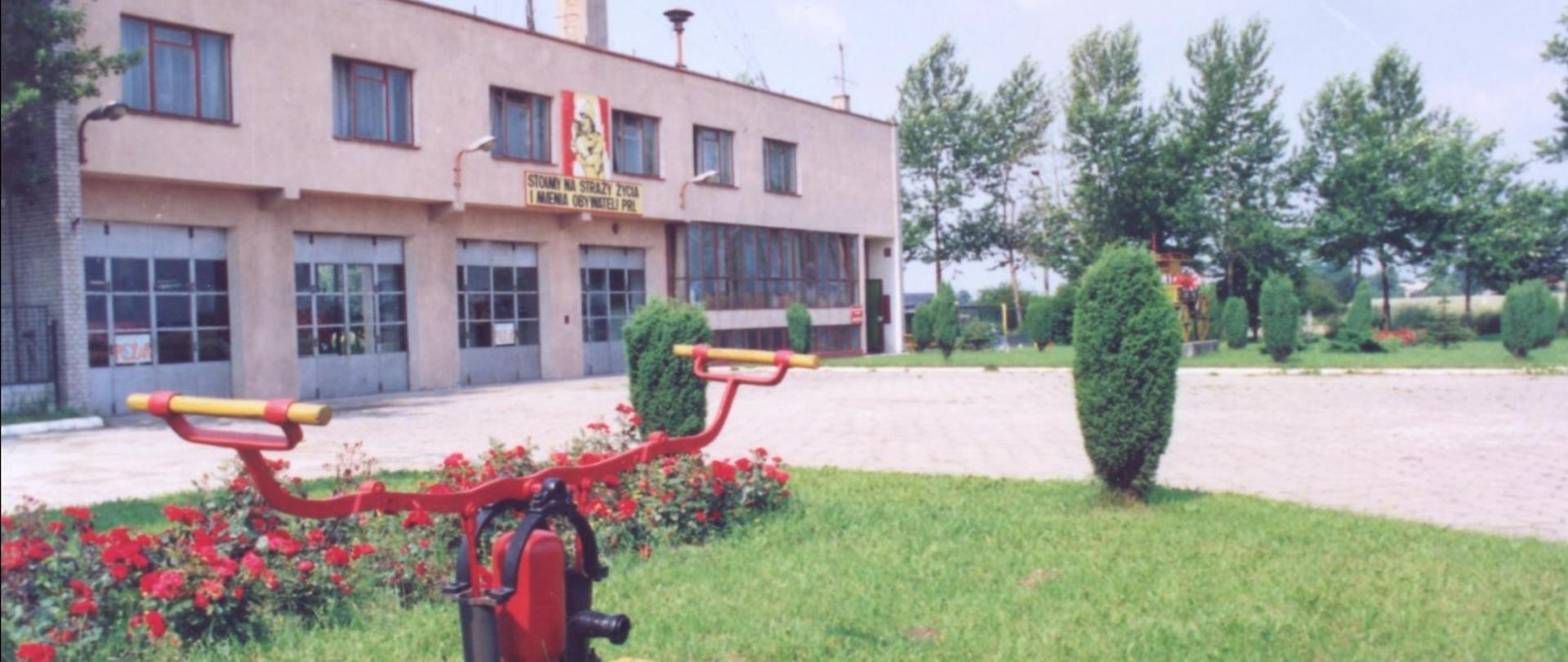 Zdjęcie przedstawia budynek Komendy Rejonowej Straży Pożarnej w Koluszkach wybudowanej w 1975r. przy ul. Słowackiego 28. Na skwerku stara pompa ręczna. 