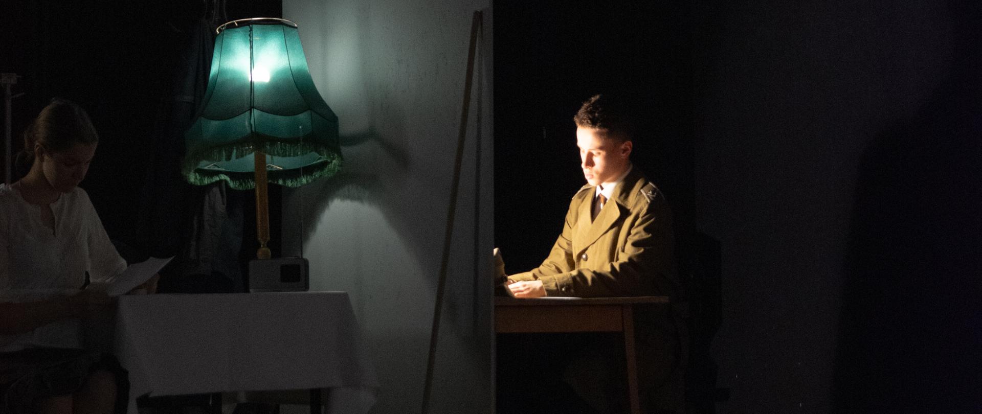 Ciemna scena teatralna, dwa stoły przedzielone ścianką. Scena oświetlona tylko lampami stojącymi na stołach. Przy stołach, po dwóch stronach ścianki, nie widząc się, siedzą kobieta i mężczyzna.