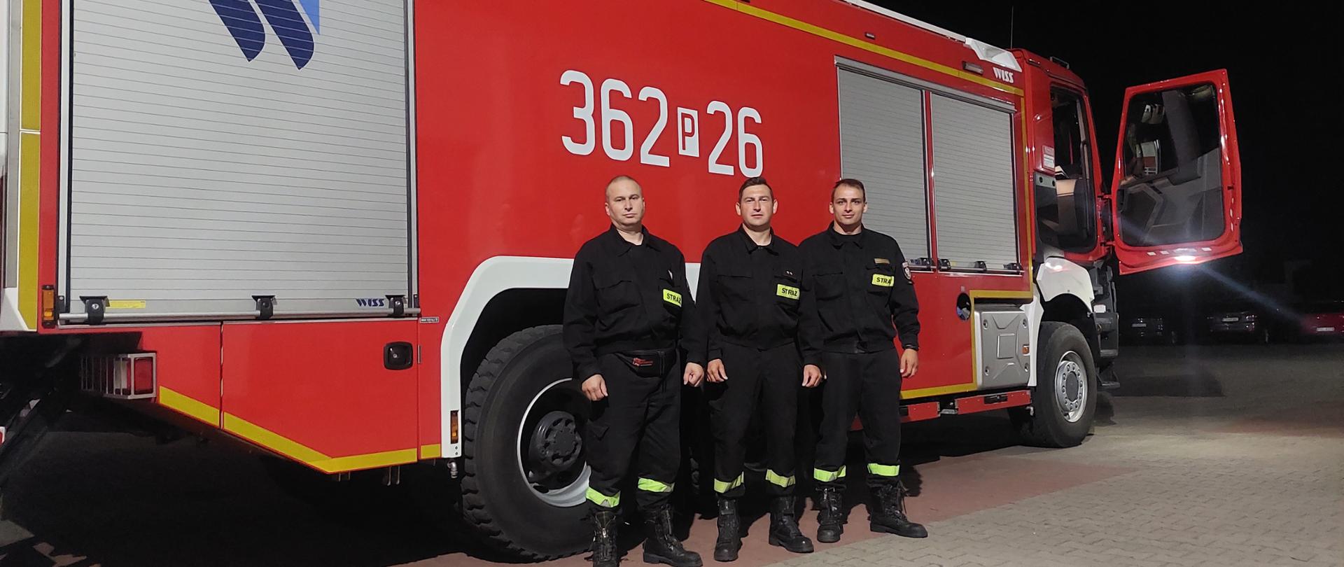 Zdjęcie przedstawia trzech strażaków stojących przed samochodem strażackim, którzy wyruszają do walki z pożarami w Grecji.
