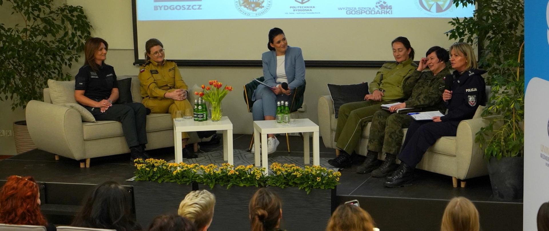 Na dwóch kanapach siedzą funkcjonariuszki służb mundurowych, każda w swoim mundurze codziennym. Po środku, na krześle siedzi moderatorka. Przed nimi dwa stoliki, kwiaty, butelki z wodą. Moderatorka trzyma w dłoni mikrofon. Z przodu donice z kwiatami. Przed tym wszystkim siedzą widzowie - widać kilkanaście osón, przodem do rozmówczyń.