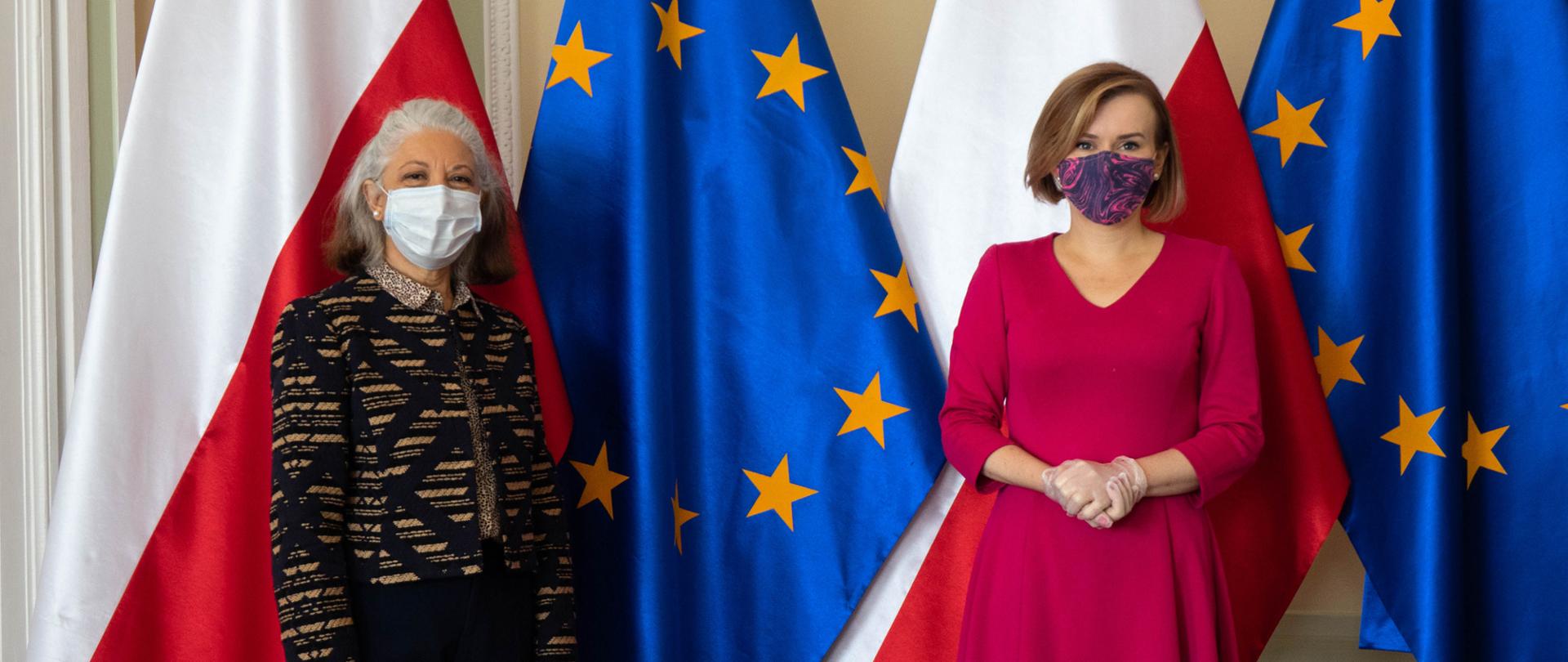 Ambasador Argentyny Pani Ana María Ramírez oraz Sekretarz Stanu Pani Anna Krupka stoją na tle flag Polski i UE. Na twarzach maseczki.