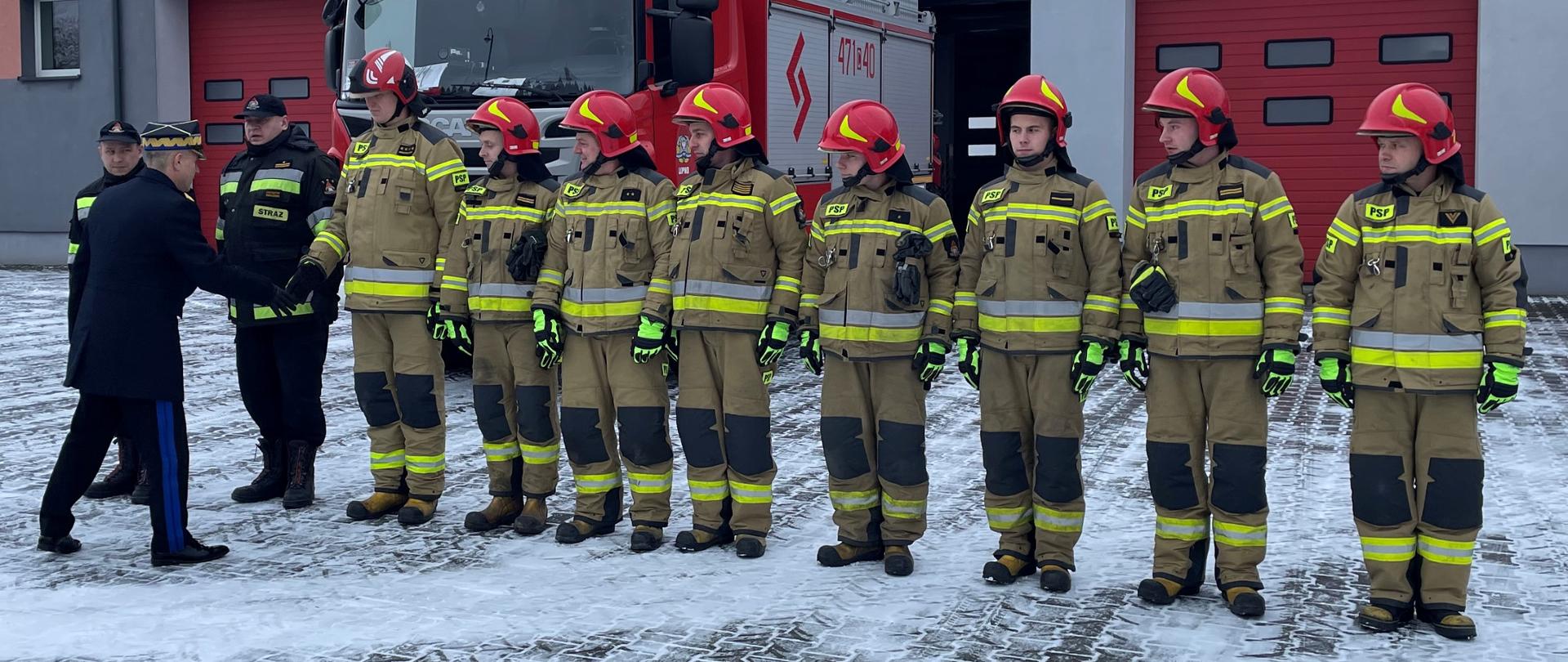 Na zdjęciu widać 10 strażaków stojących w szeregu. Komendant główny PSP wita się ze strażakami.