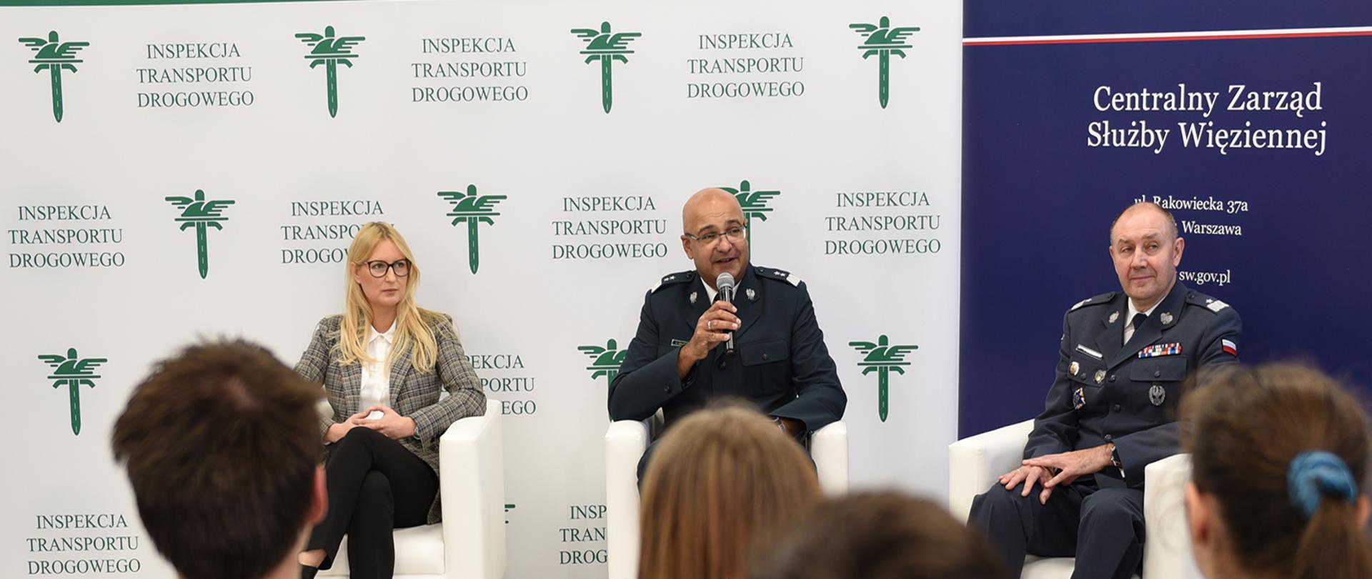 Program edukacyjny „Szanuj zasady, chroń życie” zainaugurowali Główny Inspektor Transportu Drogowego Alvin Gajadhur, Dyrektor Generalny Służby Więziennej gen. Jacek Kitliński oraz p.o. zastępca Głównego Inspektora Transportu Drogowego Klaudia Podkalicka. Na pierwszym planie młodzież siedzi na krzesłach i słucha prowadzących lekcję. Na drugim planie na tle banerów ITD i Służby Więziennej siedzą Klaudia Podkalicka, Alvin Gajadhur i Jacek Kitliński. Szef ITD Alvin Gajadhur trzyma mikrofon.