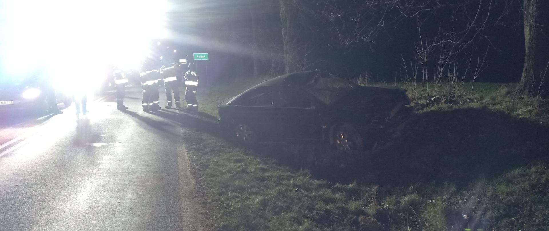 Na zdjęciu znajduje się rozbity czarny samochód osobowy. W tle znajduje się wóz strażacki oraz strażacy. Jest noc, a samochód znajduje się na zielonej trawie na poboczu jezdni.