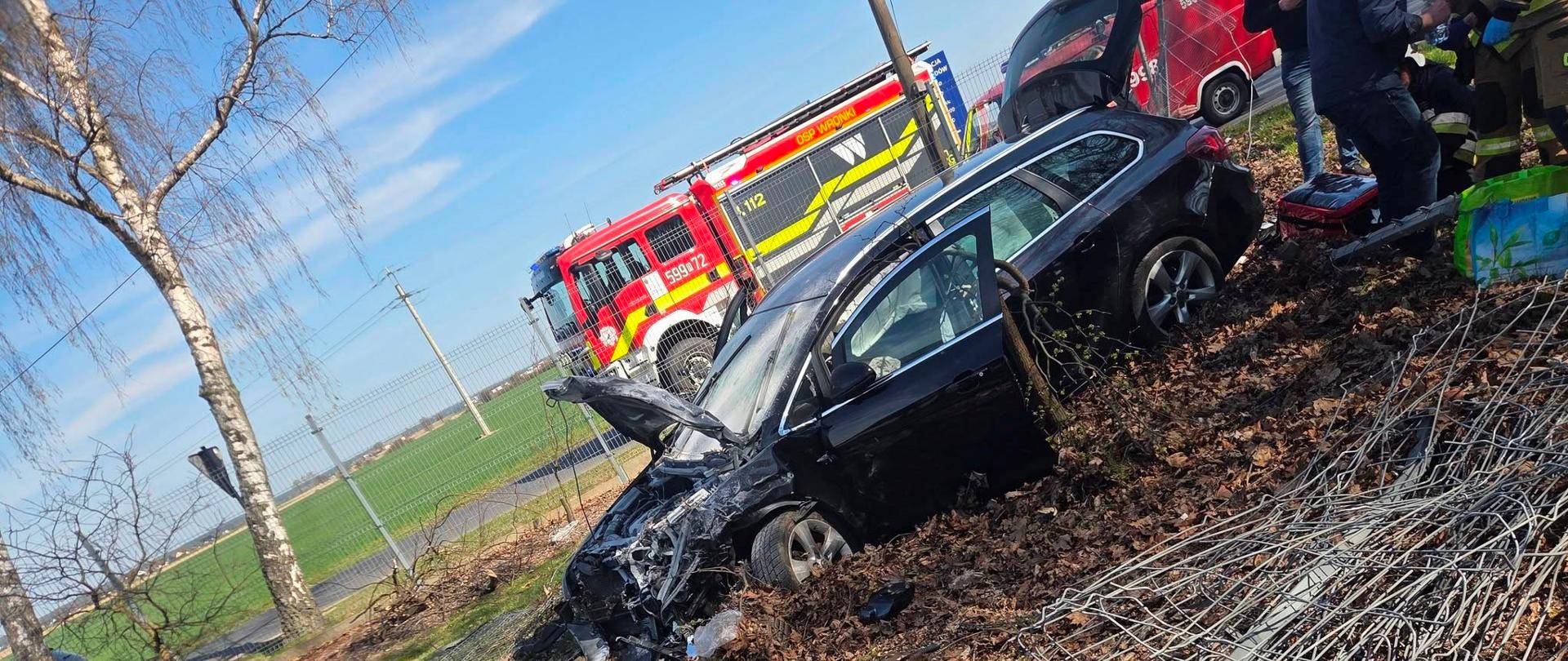 Uszkodzony pojazd na poboczu, za nim straż pożarna
