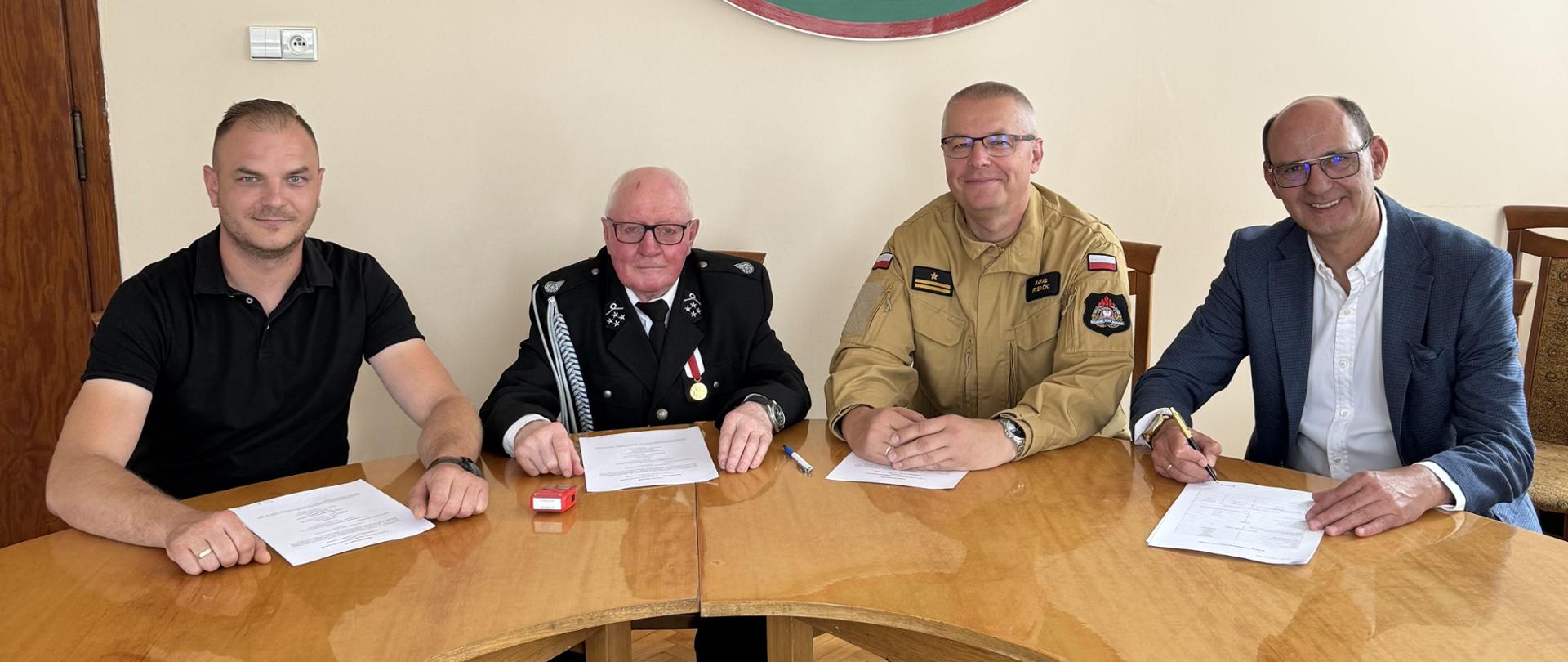Burmistrz, Prezesa, komendanta oraz naczelnika siedzących przy stole podczas podpisania porozumienia. Pozują oni do zdjęcia. Na stole leża porozumienia oraz pieczątki. Burmistrz trzyma w ręce długopis.