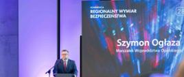 Na zdjęciu za mównicą stoi marszałek Szymon Ogłaza. W tle ścianka multimedialna. Na dole zdjęcia widać siedzących tyłem innych uczestników konferencji. 
