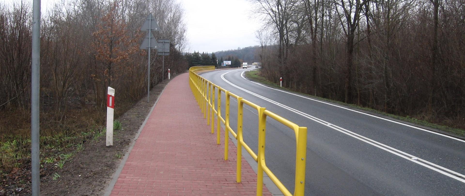 Chodnik wzdłuż drogi krajowej - chodnik z czerwonej kostki oddzielony od jezdni żółtymi barierkami 
