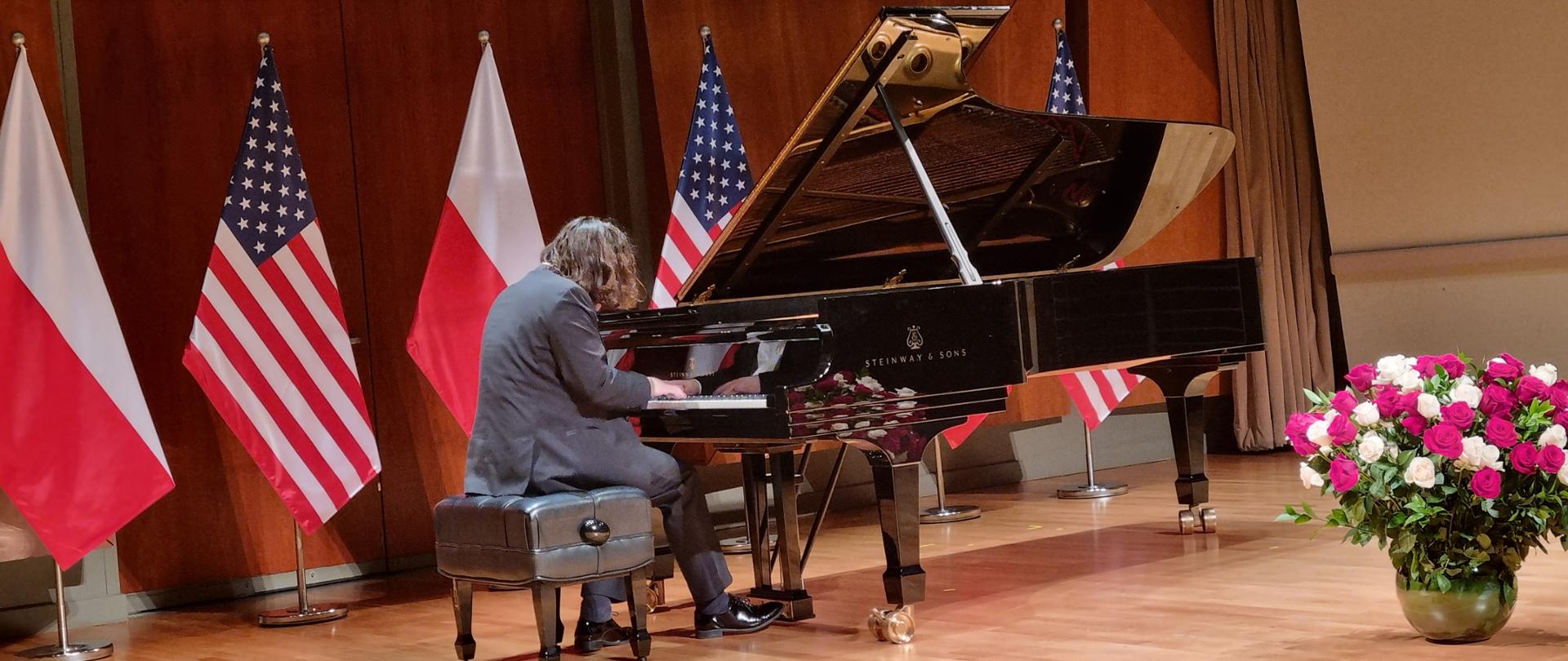 Recital fortepianowy Jakuba Kuszlika z okazji Narodowego Święta Niepodległości Polski. Pianista wykonuje utwory polskich kompozytorów na czarnym królu na scenie w Colburn School w Los Angeles. W tle flagi Polski i Stanów Zjednoczonych. Przed fortepianem stoi biało-czerwony bukiet róż. 