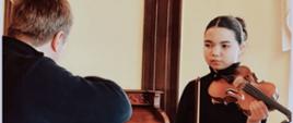 Zdjęcie kolorowe. Dziewczyna ubrana w czarny golf, trzymająca skrzypce i smyczek, patrząca w stronę mężczyzny odwróconego plecami. Mężczyzna w ciemnej bluzie, z widocznym profilem głowy i prawym ramieniem. W tle jasna ściana, ciemnobrązowy drewniany filar oraz rzeźbiony mebel przypominający biurko lub pianino. Jasne, ciepłe oświetlenie wnętrza.