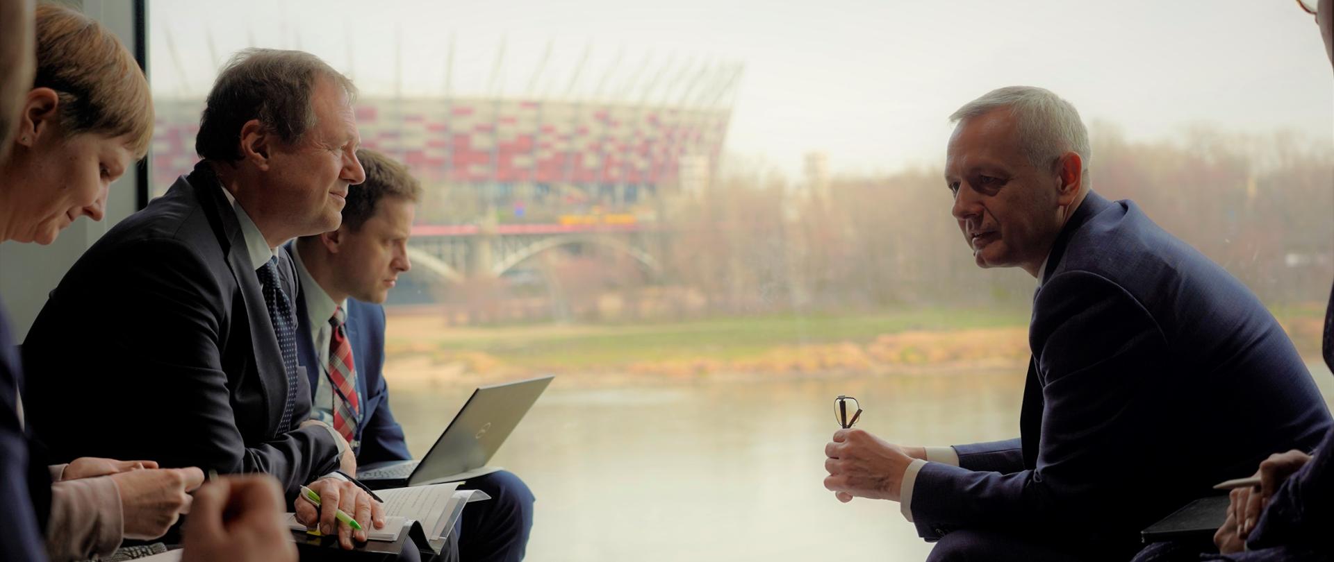 Kilka osób siedzi przy małym stoli i rozmawia. Wśród nich minister Marek Zagórski. W tle za oknem widać Stadion Narodowy w Warszawie.