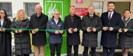 Trzy kobiety i pięciu mężczyzn stoją przed budynkiem i trzymają w rękach zieloną wstęgę i nożyczki. Wszyscy uśmiechają się do zdjęcia.