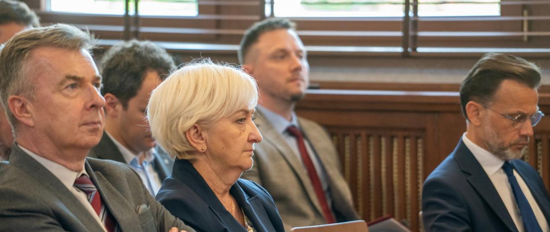 Na krzesłach siedzą ludzie w garniturach. W centralnej części zdjęcia starsza kobieta o siwych włosach, ubrana w granatową garsonkę. 